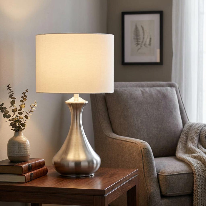 Modern satin silver touch lamp with a white fabric shade lit on a wooden side table next to an armchair.