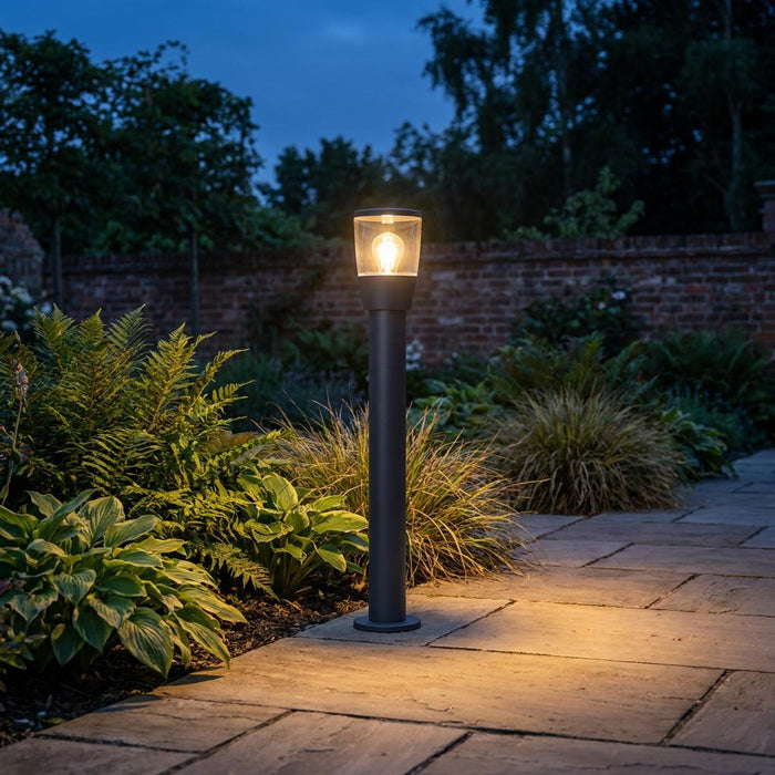 Warmly lit black bollard light beside a stone path in a garden at dusk.