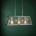 Chrome three-light lantern bar with clear glass panels and lit bulbs against a dark green wall.