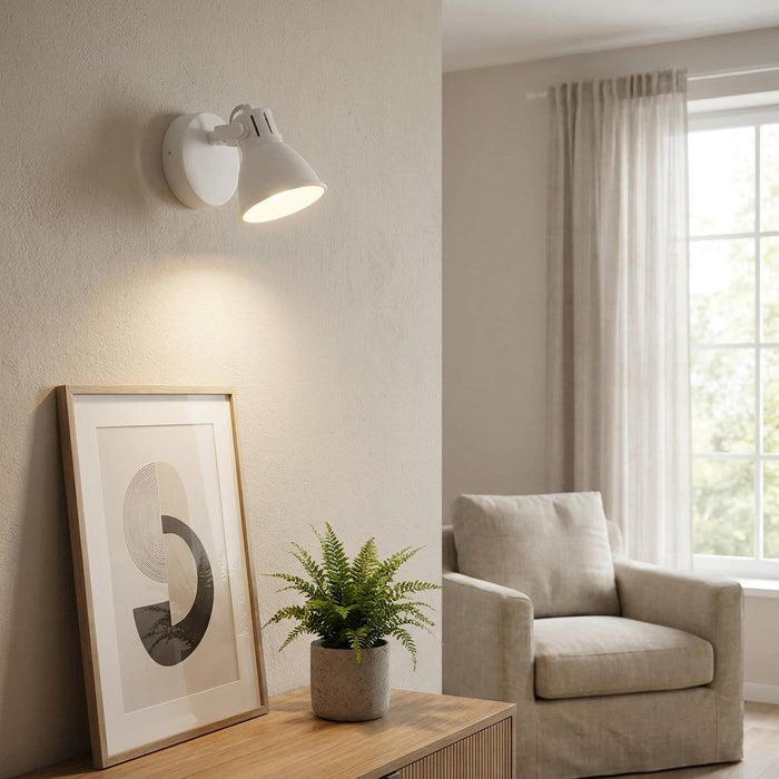 White wall spotlight illuminating framed art and a potted fern on a wooden sideboard in a living room.