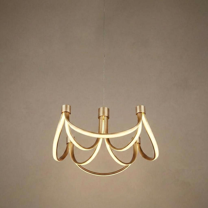 A gold 8-arm LED pendant light with looping ribbon-like arms hanging against a textured neutral wall.