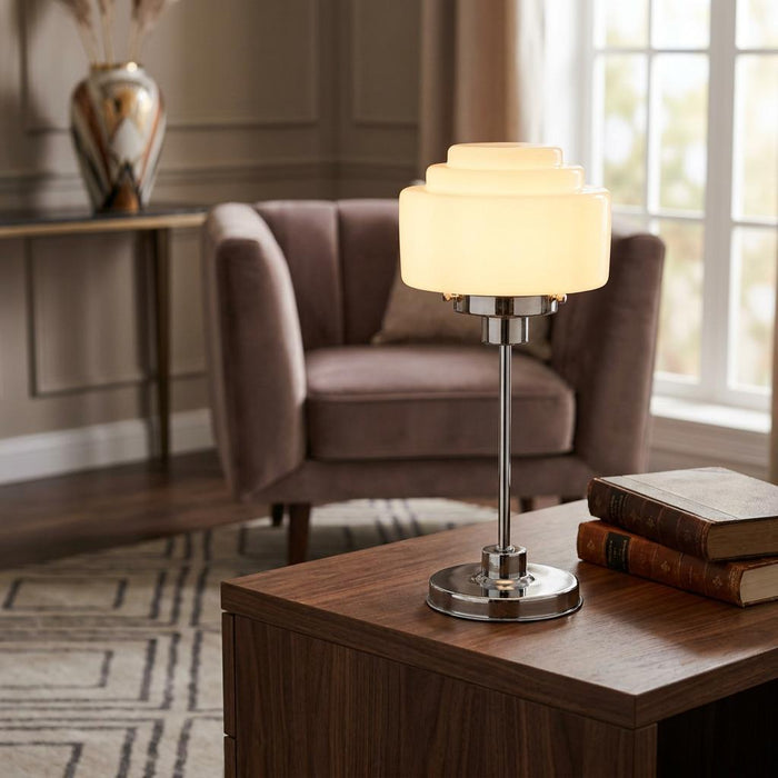 Small opal glass school lamp with a tiered shade and chrome base on a wooden table in a cozy living room.