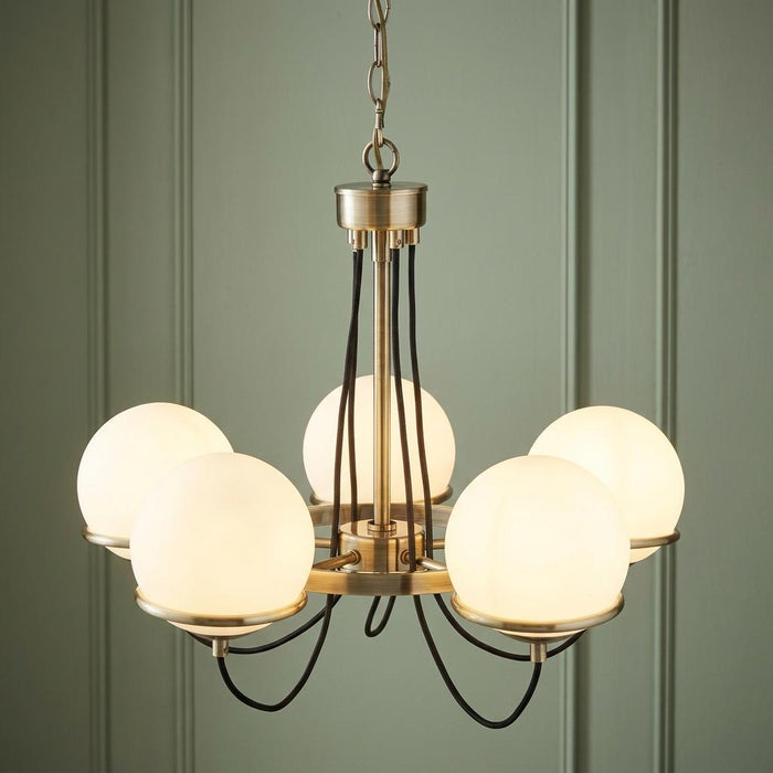Five-light brass chandelier with white globe shades and braided cables against a green paneled wall.