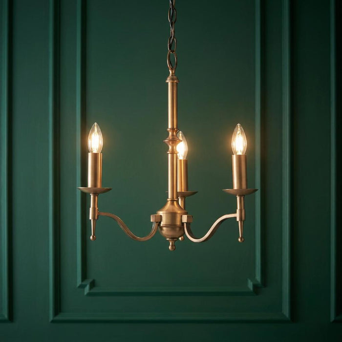Antique brass three-light chandelier with lit candle bulbs against a dark green paneled wall.