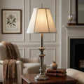Elegant antique nickel table lamp with a cream paneled shade lit on a wooden side table in a cozy living room.