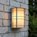 Stockholm galvanised steel outdoor wall light with a warm glow mounted on a white brick wall.