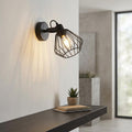 Black wire cage wall spotlight illuminated on a white wall above a shelf with a small plant.