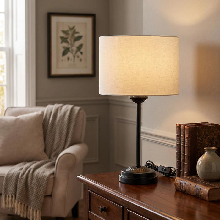 Black slim table lamp with a lit cream drum shade on a wooden sideboard in a warm living room.