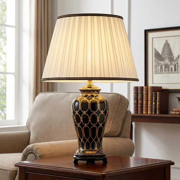 Taipei ceramic table lamp with an ornate black and gold base and ivory pleated shade on a wooden side table.