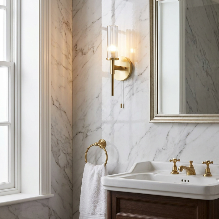 Illuminated brass wall light with a ribbed glass shade mounted on a marble bathroom wall next to a mirror.