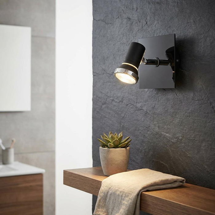 Black and chrome wall spotlight mounted on a textured wall, illuminating a succulent and towel on a wooden shelf.