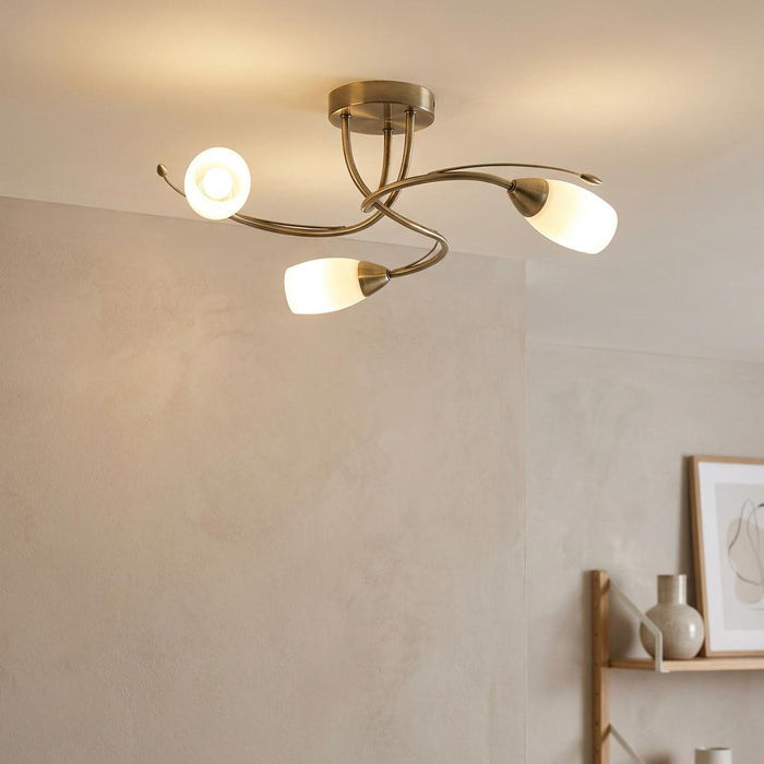 Antique brass flush ceiling light with three curved arms and frosted glass shades glowing in a neutral room.