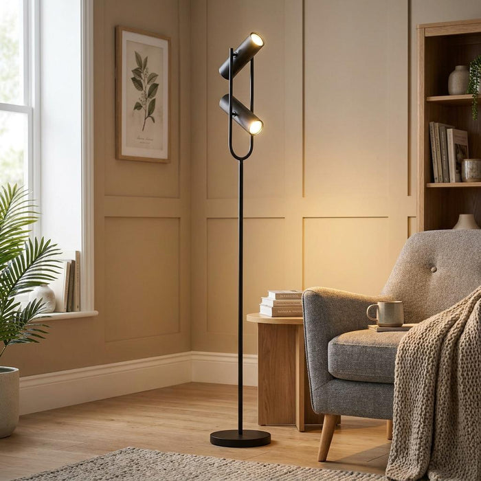 Modern black floor lamp with two adjustable spotlights illuminating a neutral living room with an armchair.