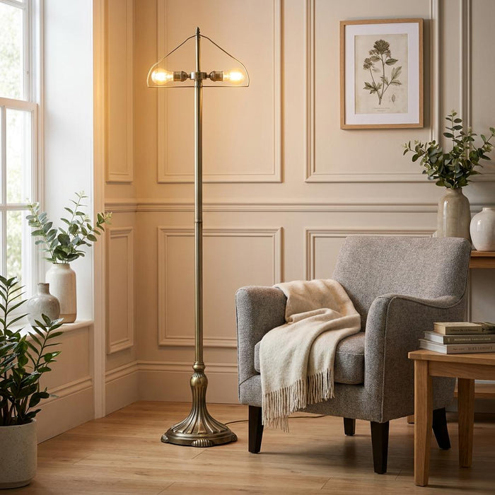 Antique brass floor lamp base with two glowing bulbs beside a grey armchair in a neutral-toned living room.