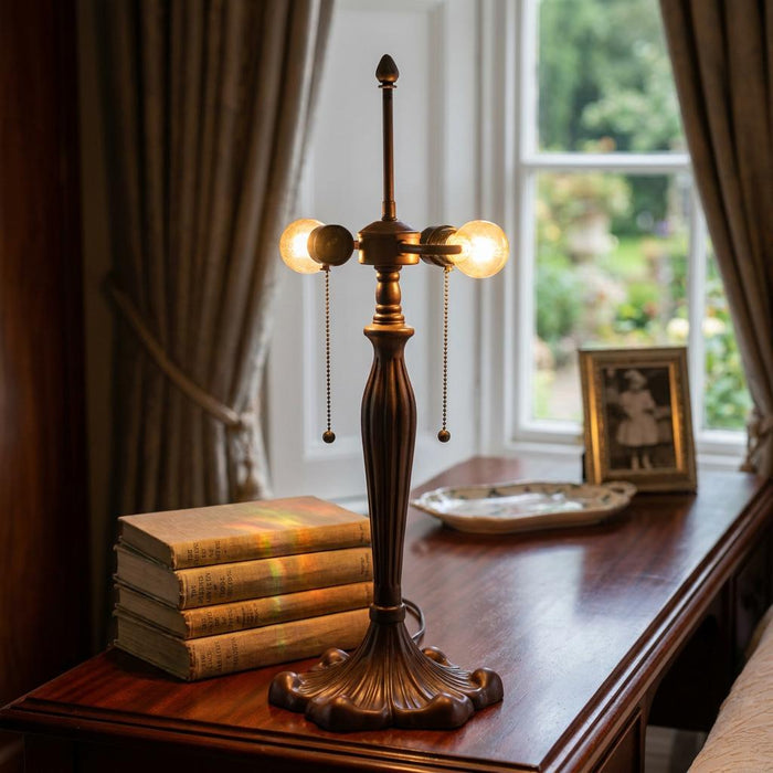 Bronze double bulb lamp base with pull chains on a dark wood desk next to a stack of books.