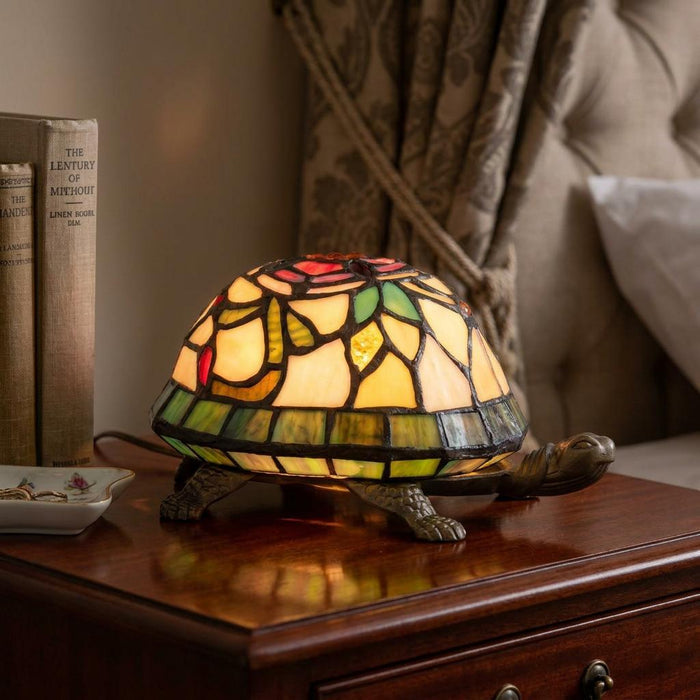 Illuminated Tiffany tortoise table lamp with a colorful stained glass shell on a dark wooden nightstand.