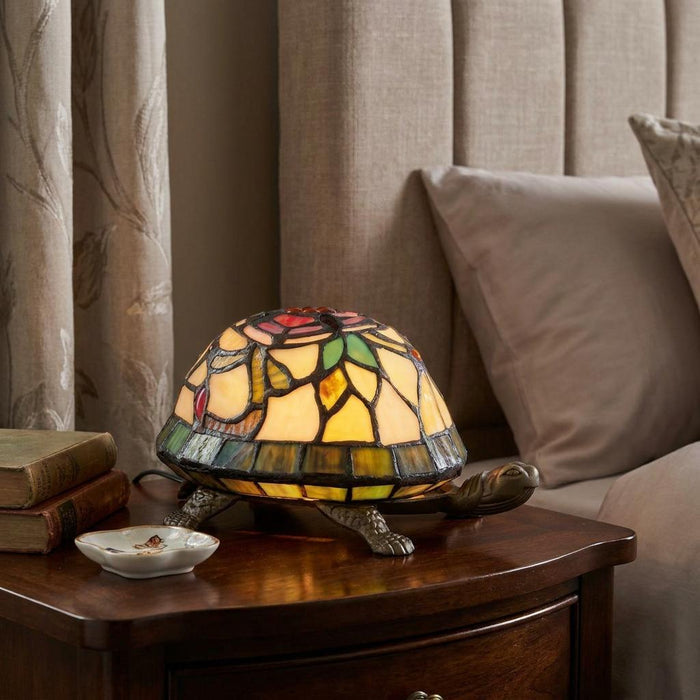 Lit Tiffany-style tortoise lamp with a colorful stained glass shell on a wooden bedside table.