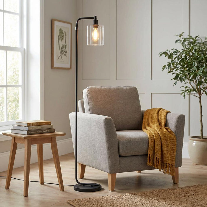 Toledo Black Floor Lamp with a clear glass shade stands next to a grey armchair with a yellow throw in a living room.