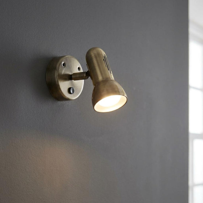 Antique brass adjustable wall spotlight illuminated against a dark grey wall.
