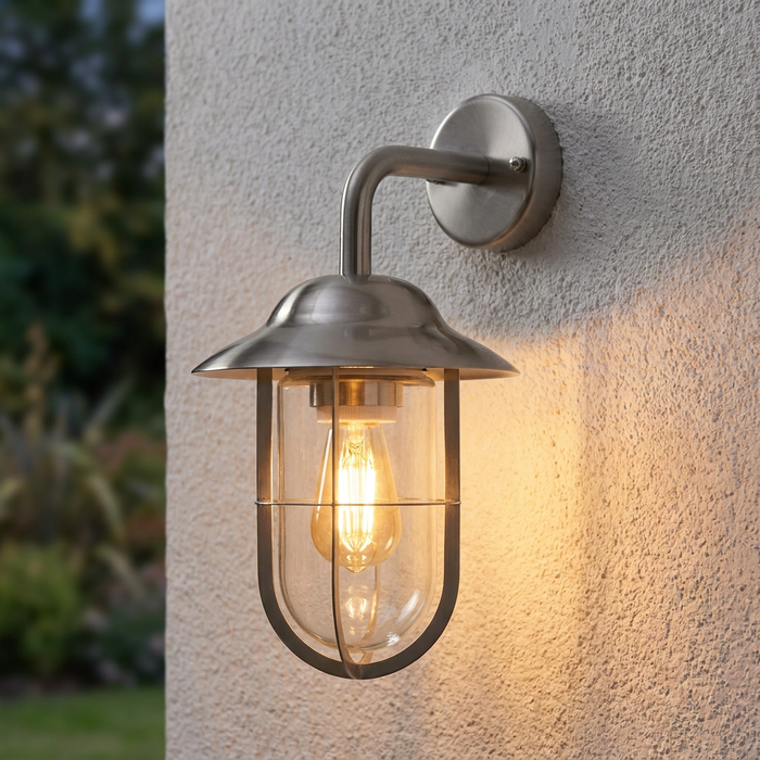 Lit satin silver lantern wall light with a glass shade and protective cage on a white exterior wall.