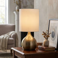 Touch table lamp with an antique brass teardrop base and white drum shade on a wooden table in a cozy living room.