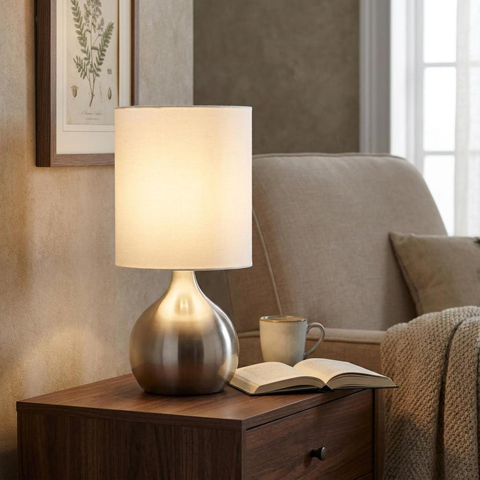 Illuminated satin silver touch lamp with a white drum shade on a wooden side table in a cozy living room.