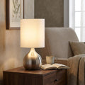 Illuminated satin silver touch lamp with a white drum shade on a wooden side table in a cozy living room.