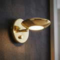 Polished brass wall light with a saucer-shaped adjustable head and dimmer switch on a dark textured wall.