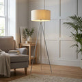 Tri chrome tripod floor lamp with a cream drum shade standing in a modern living room next to an armchair.