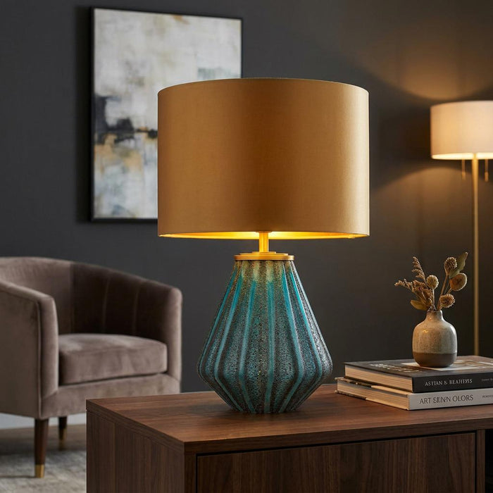 Turquoise ribbed glass table lamp with a gold drum shade on a wooden table in a modern living room setting.