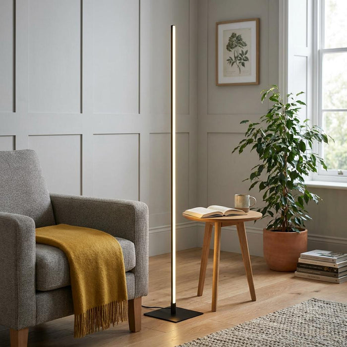 Minimalist black LED floor lamp illuminating a cozy living room corner with a grey armchair and wooden side table.