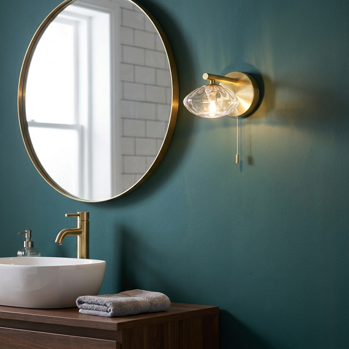 Brass wall light with textured glass shade and pull cord, lit on a dark teal wall next to a round vanity mirror.
