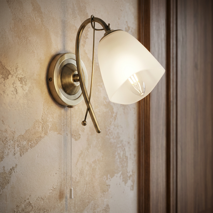 Antique brass wall light with a frosted glass shade and pull cord mounted on a textured wall.