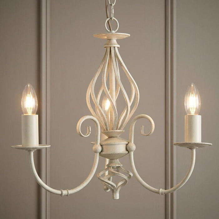 Tuscany ivory finish 3 light chandelier featuring elegant scrollwork and lit candle bulbs against a paneled wall.