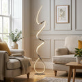 Spiral-shaped chrome LED floor lamp standing between two beige armchairs in a light-filled living room.