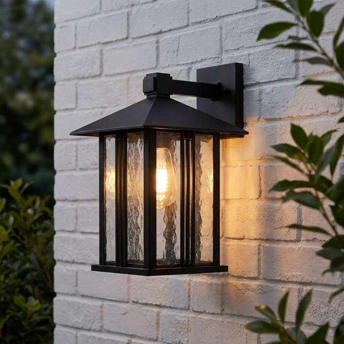 Illuminated black lantern wall light with textured water glass panels mounted on a white brick wall.