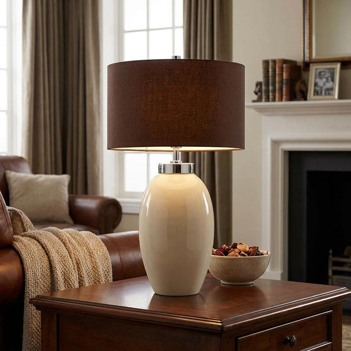 Victor table lamp with a cream ceramic oval base and brown fabric shade on a wooden table in a living room.
