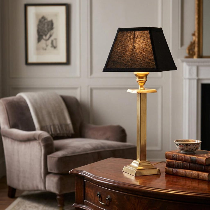 Solid brass table lamp with a black tapered shade on a wooden side table in a traditional living room.