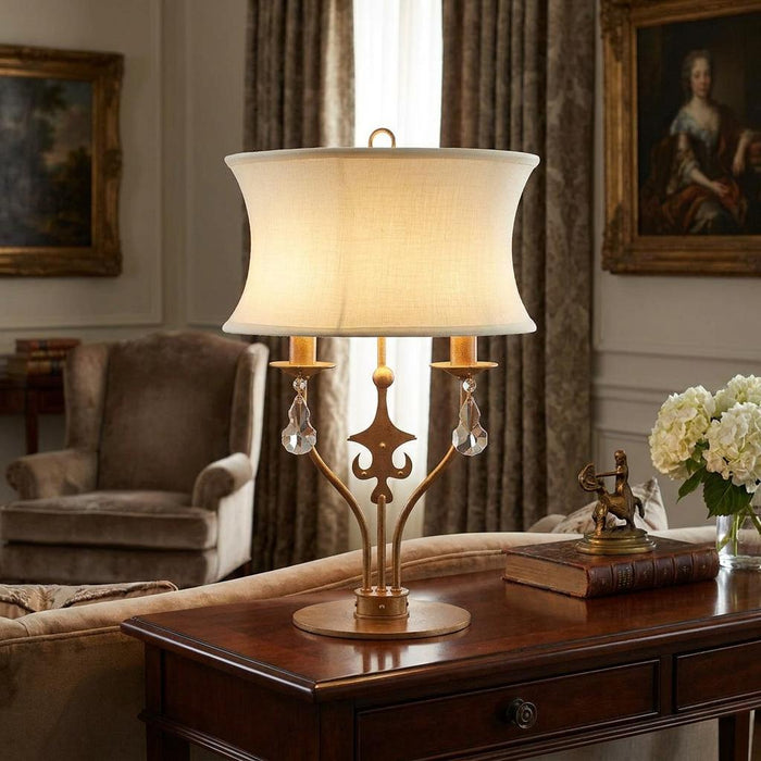 Gold table lamp with a curved cream shade, crystal drops, and fleur-de-lis motif on a wooden table.