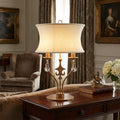 Gold table lamp with a curved cream shade, crystal drops, and fleur-de-lis motif on a wooden table.