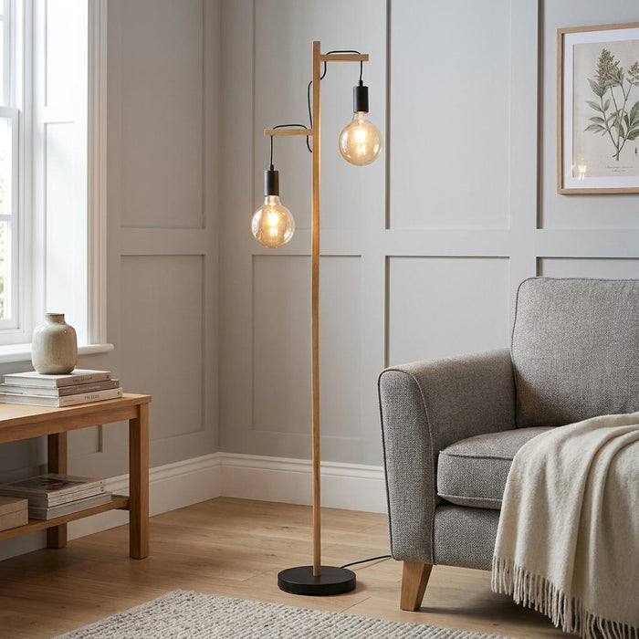 Two-light ash wood floor lamp with large globe bulbs next to a grey armchair in a contemporary living room.