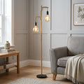 Two-light ash wood floor lamp with large globe bulbs next to a grey armchair in a contemporary living room.