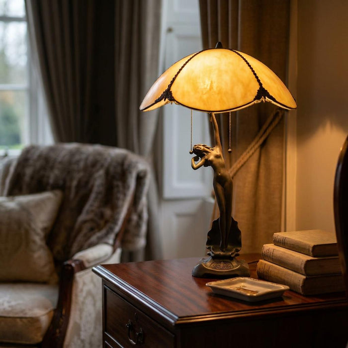 Tiffany table lamp featuring a bronze female figure base and amber art glass shade on a dark wood nightstand.