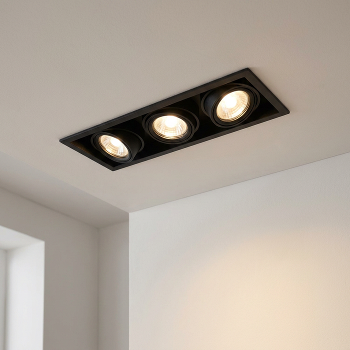 A black triple recessed spotlight fixture mounted on a white ceiling, casting warm light on the wall below.