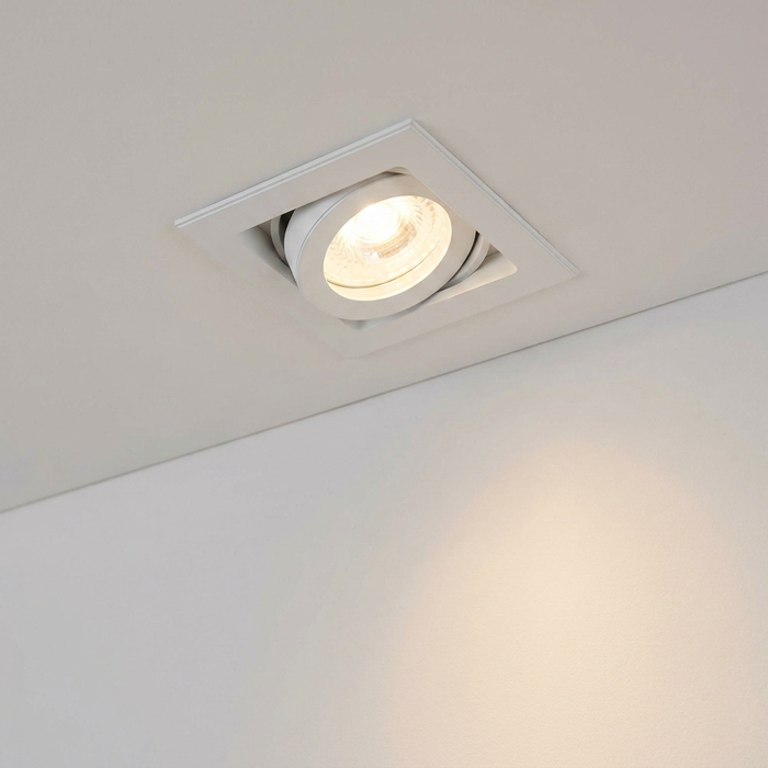 White square recessed adjustable spotlight tilted and illuminated in a white ceiling.