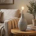 Textured grey ceramic table lamp base on a wooden side table with a lit bulb in a living room setting.