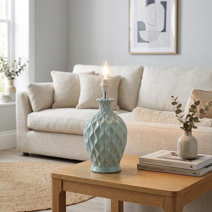 Light blue textured ceramic lamp base with a glowing bulb on a wooden table in a brightly lit living room.
