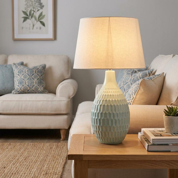 Light blue textured ceramic table lamp with a cream shade lit on a wooden side table in a living room.