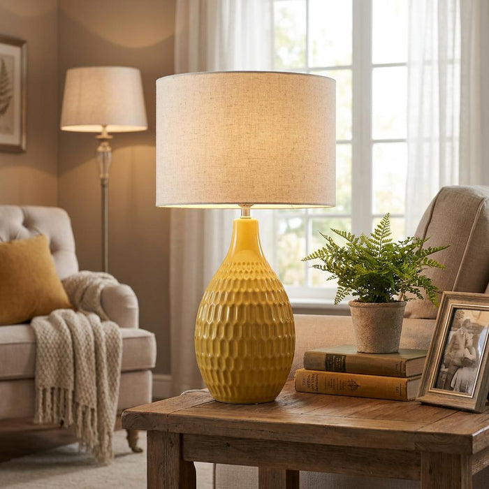 Yellow textured ceramic table lamp with a beige shade on a rustic wooden side table in a warm living room.