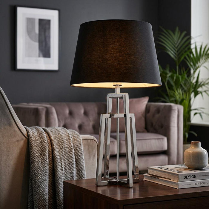 Chrome crossed frame table lamp with a black fabric shade illuminated on a wooden table in a contemporary living room.